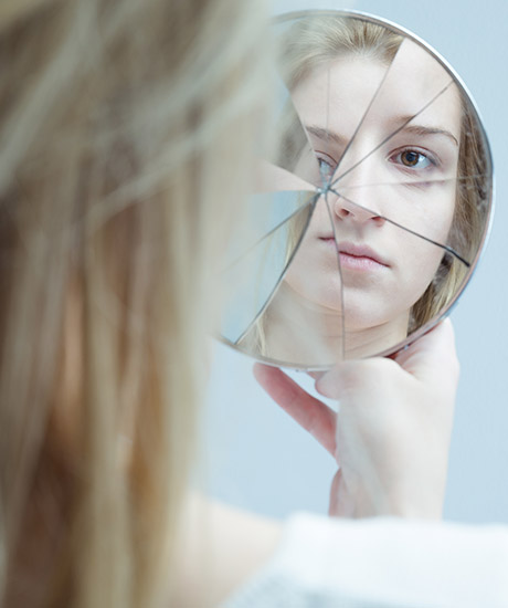 Het spiegelbeeld van het gezicht van een jonge vrouw, gezien over haar schouder. De spiegel is gebroken in kleine stukjes.